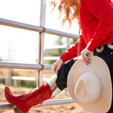 Person wearing red cowboy boots and a red sweater, holding a white cowboy hat showing off Lucky Chuck Boot Socks.