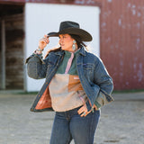 Woman wearing a sts racnhwear denim jacket, sts sherpa, and jeans with a black cowboy hat in front of a red barn.
