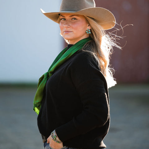 Woman wearing a cowboy hat, black sweater, and green silk wild rag in an outdoor setting